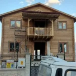 Balkonu olan önünde aracı ve girişi gözüken iki katlı ağaç tomruk ev