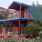 Kızıl ile mavinin tonları ile iki katlı altta veranda veranda üstünde balkonu olan dik çatılı yeşil şıngıl olan ahşap ev görseli